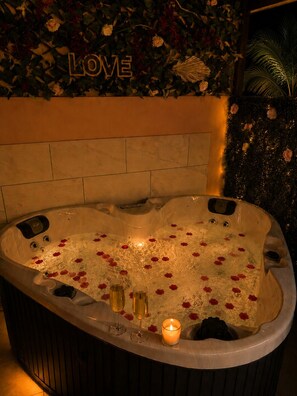Ein Whirlpool mit Rosenblättern und Kerzen, umgeben von einer Wand mit einem 'LOVE'-Schild und Blumendekorationen.