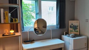 Ein Schlafzimmer mit einem Bett, einem Schreibtisch mit Spiegel, einem kleinen Kühlschrank und einem Fenster mit Vorhängen.