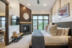 A modern bedroom with a large bed, a fireplace, a view of the outdoors, and a framed picture on the wall. - Dogwood and Stone Luxury 2BR Mountain Escape 3K (Park City)