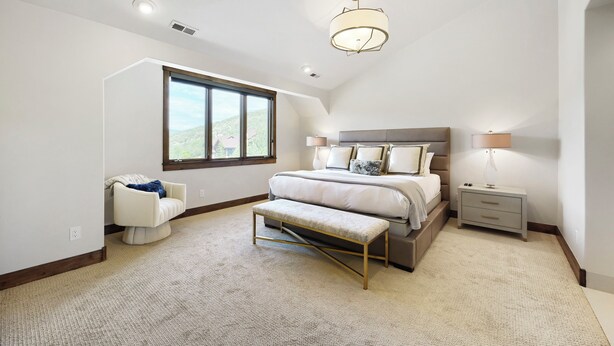 A bedroom with a large bed, a bench, a nightstand, a lamp, and a window with a view of a hill. - 1491 April Mountain Drive (Park City)