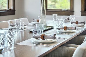 Una mesa de comedor dispuesta con cristalería transparente, platos y cubiertos, con un florero como centro de mesa y vistas al exterior.