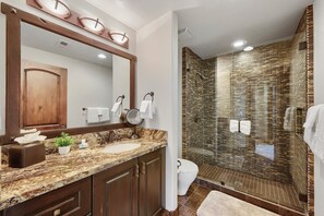 Un baño con encimera de mármol, un mueble de madera, un espejo, un lavabo, un inodoro y una ducha con puerta de cristal.