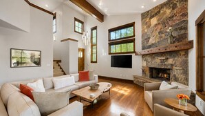 A spacious living room with a stone fireplace, a large TV, and a view of the outdoors through multiple windows. - 1491 April Mountain Drive (Park City)