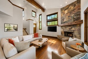 A spacious living room with a stone fireplace, a large TV, and a view of the outdoors through multiple windows. - 1491 April Mountain Drive (Park City)