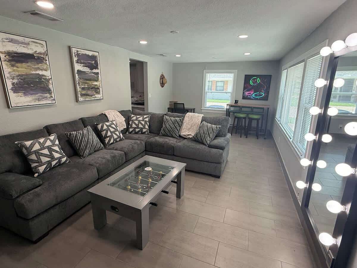 A living room with a sectional sofa, a glass-top coffee table, and a wall-mounted mirror with lights.