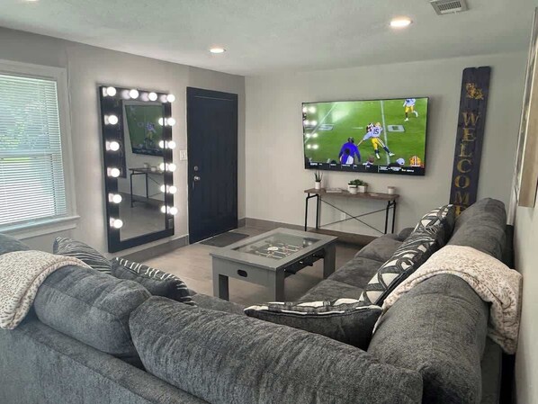 A living room with a gray sofa, a glass-top coffee table, a TV mounted on the wall, and a mirror with lights.