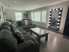 A living room with a sectional sofa, a glass-top coffee table, and a large mirror with lights.