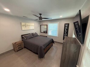 Ein Schlafzimmer mit einem Bett, einem Nachttisch, einem Deckenventilator, einem an der Wand montierten Fernseher und einem Fenster mit Jalousien.