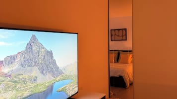 Een moderne woonkamer met een flatscreen televisie op een witte standaard, een houten vloer en een gezellig bed met een witte deken.