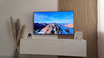 Een moderne woonkamer met een flatscreen televisie aan een houten wand, een witte kast eronder en een ronde salontafel met afstandsbedieningen en tijdschriften.