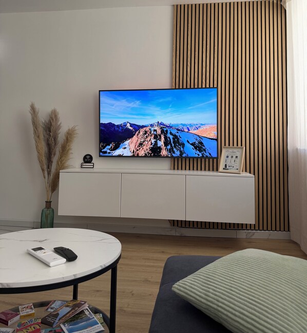 Una sala de estar moderna con un televisor de pantalla plana montado en una pared de madera, un mueble blanco debajo y una mesa redonda de centro con controles remotos y revistas.