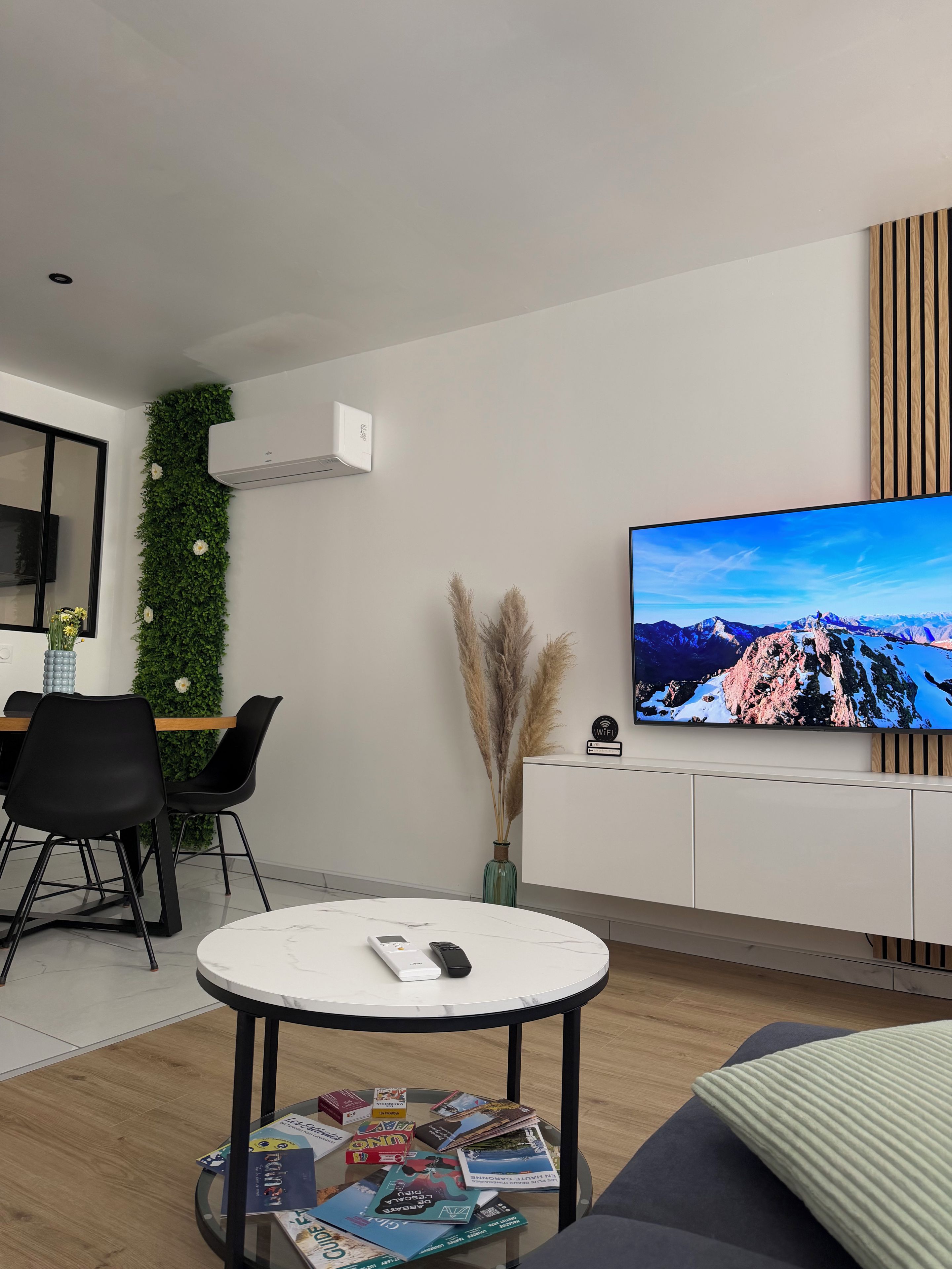 A modern living room with a flat-screen TV, a round coffee table with magazines, a sofa, a dining area with chairs, and a wall-mounted air conditioner.