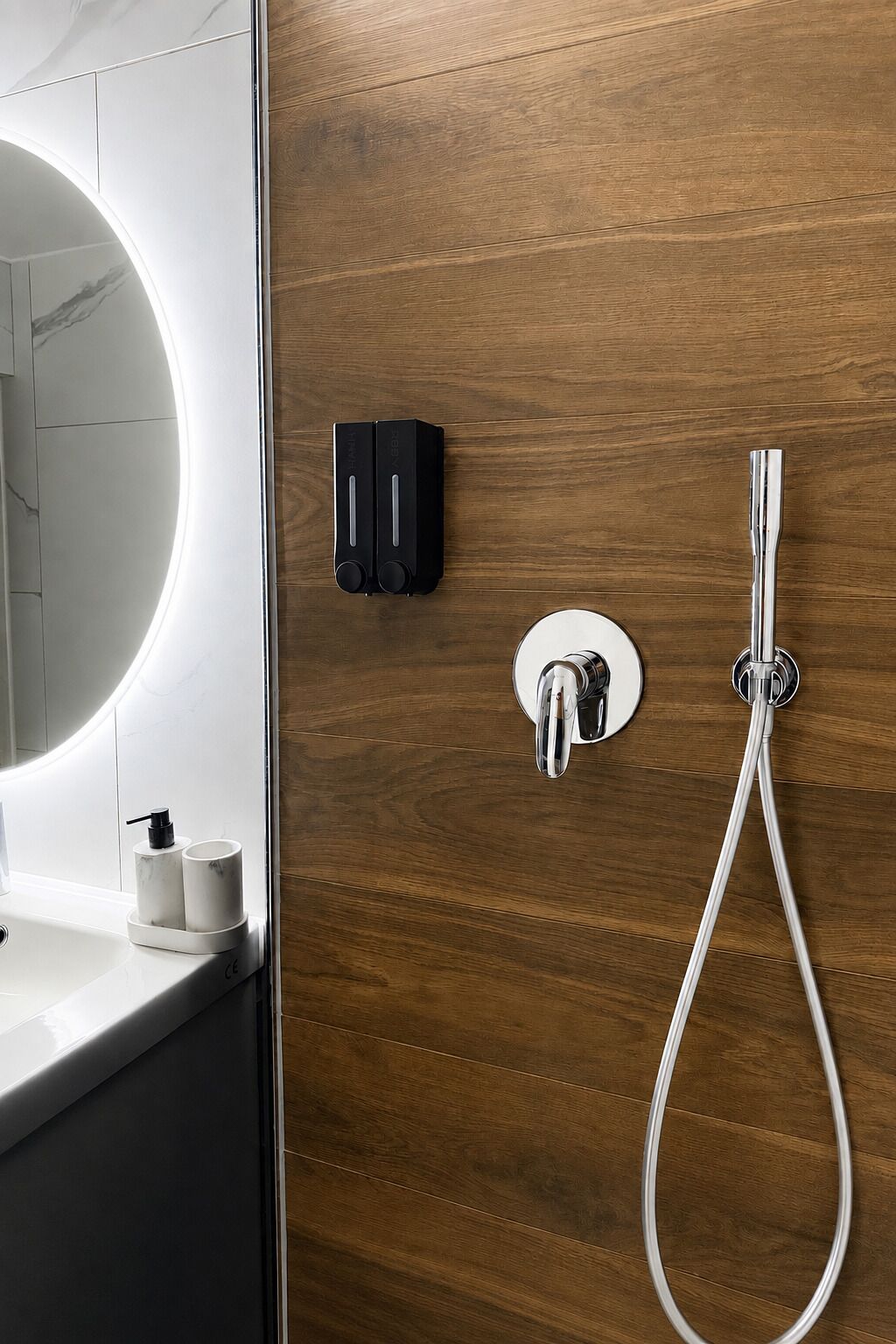 Ein Badezimmer mit Holzwand, Waschbecken in Weiß, Spiegel, Duschkopf und Seifenspender.