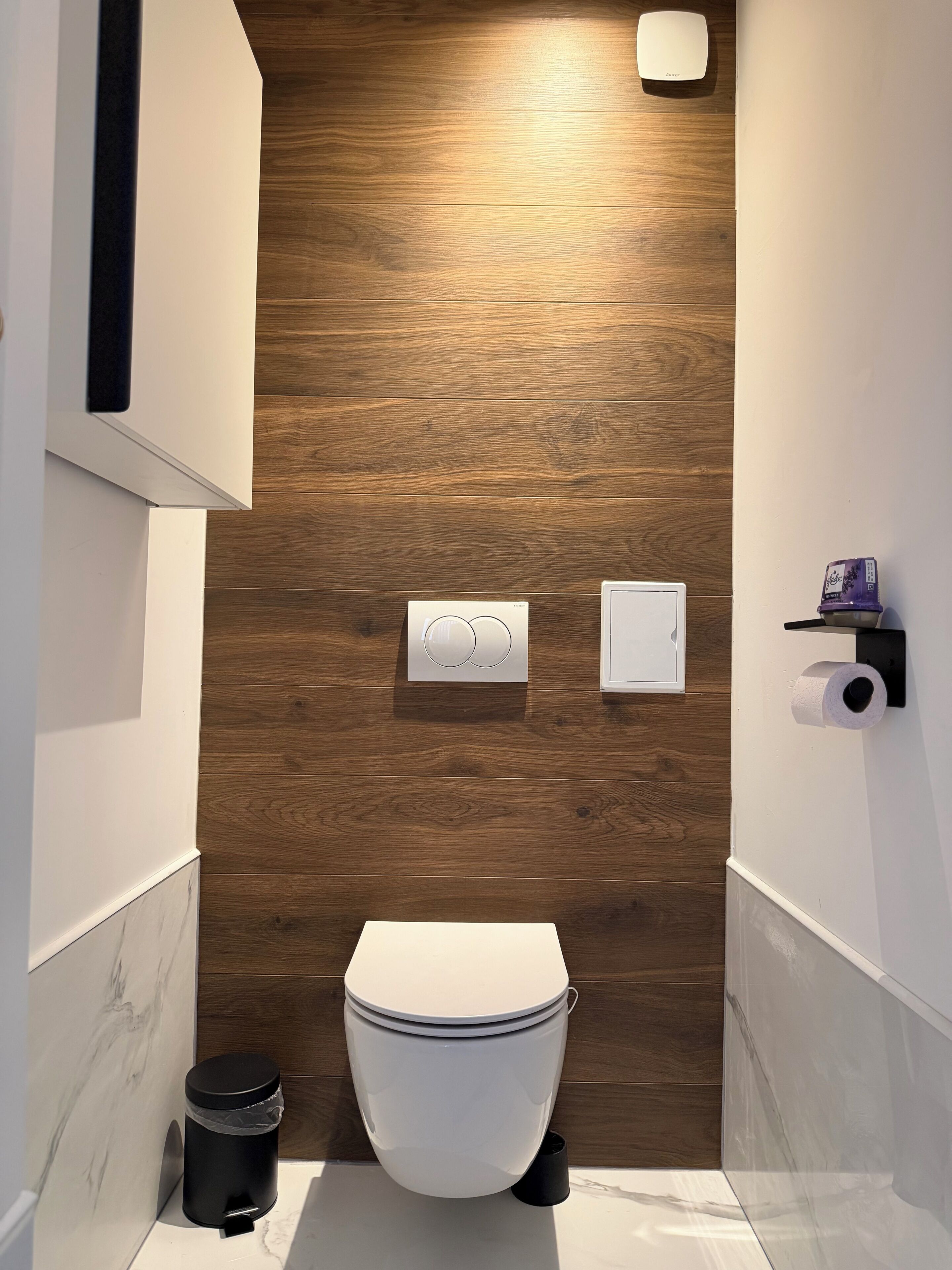 A modern bathroom with a wooden floor, a white toilet, a black trash bin, and a toilet paper holder.