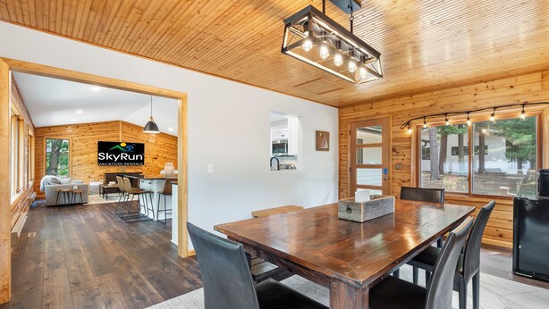 Ett rymligt matområde med ett träbord och stolar, ett kök som syns genom en öppen dörr och en logotyp för SkyRun Vacation Rentals på väggen.