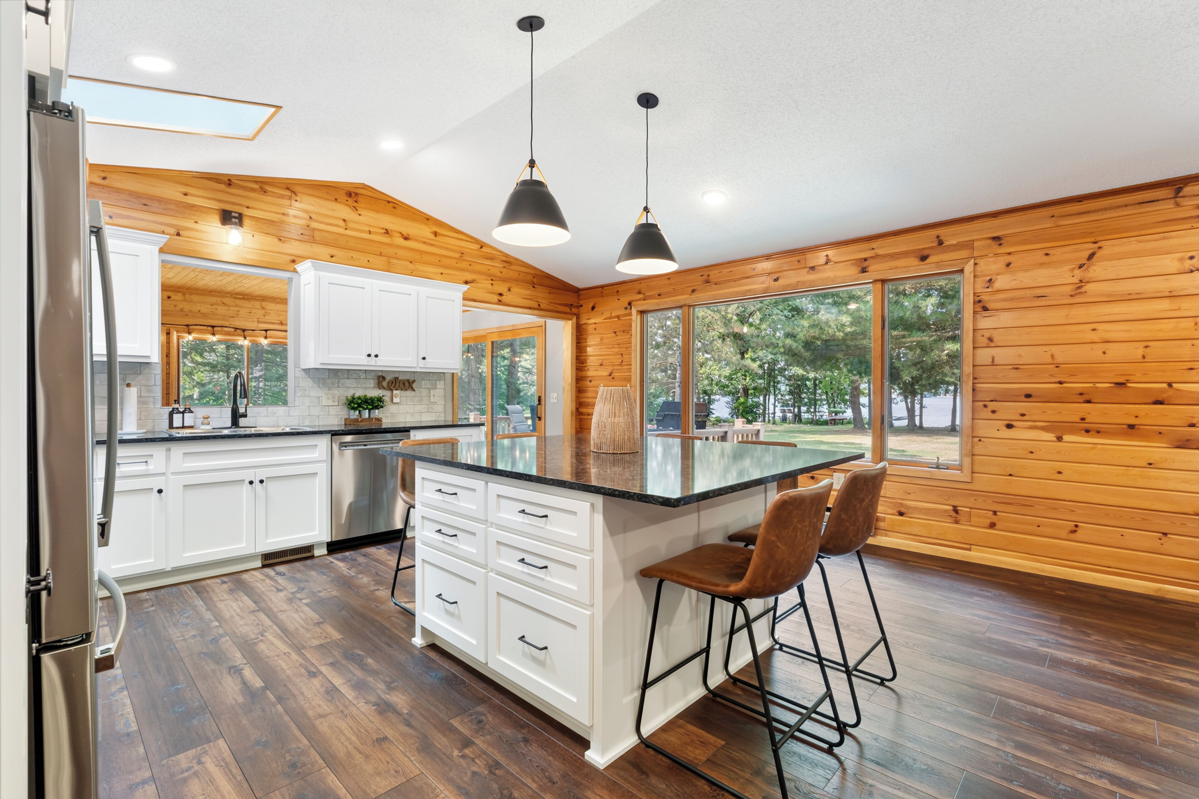 A modern kitchen with wooden walls, white cabinets, a large island with bar stools, and a view of the outdoors.