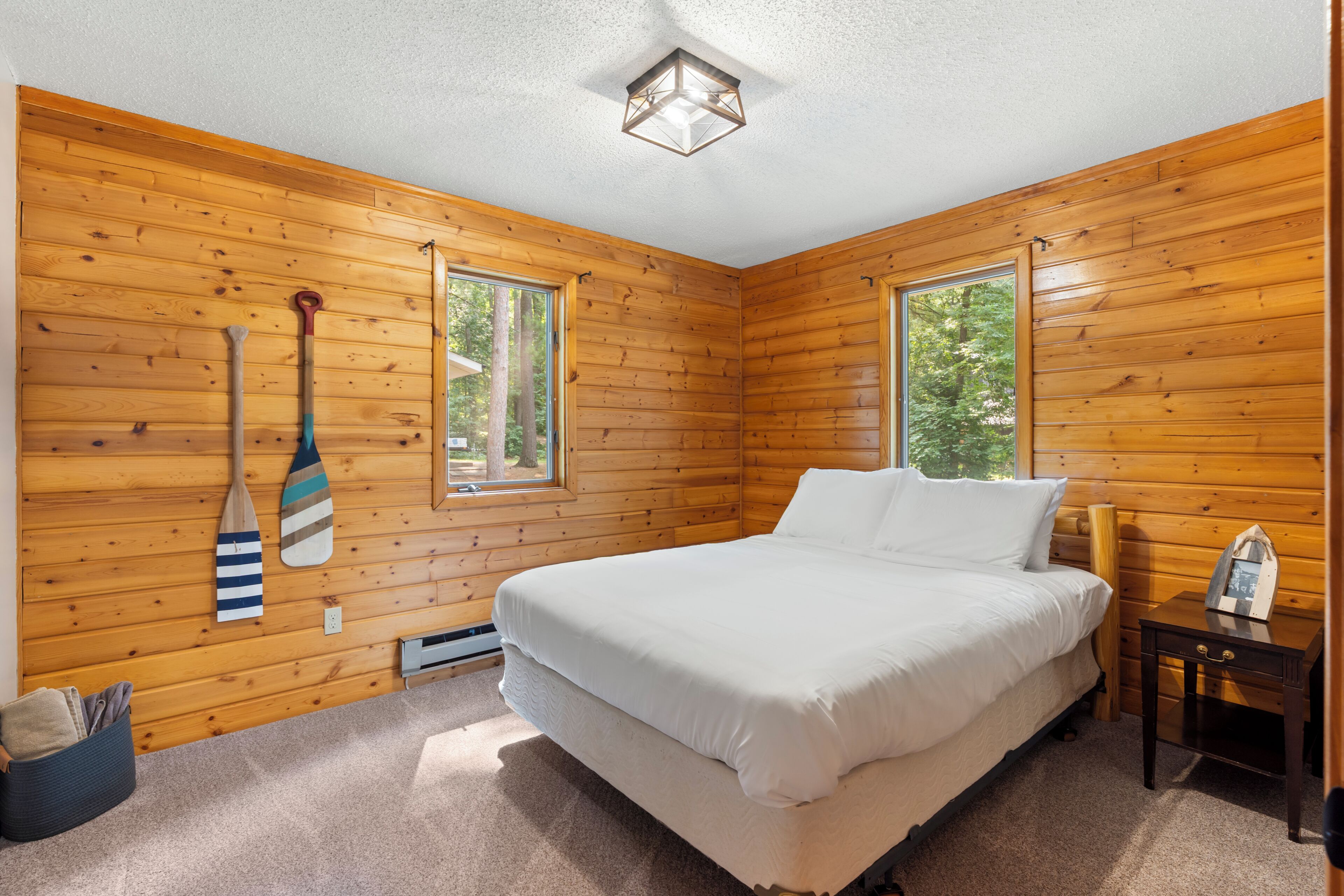 A bedroom with wooden walls, a bed, a nightstand, and two windows.
