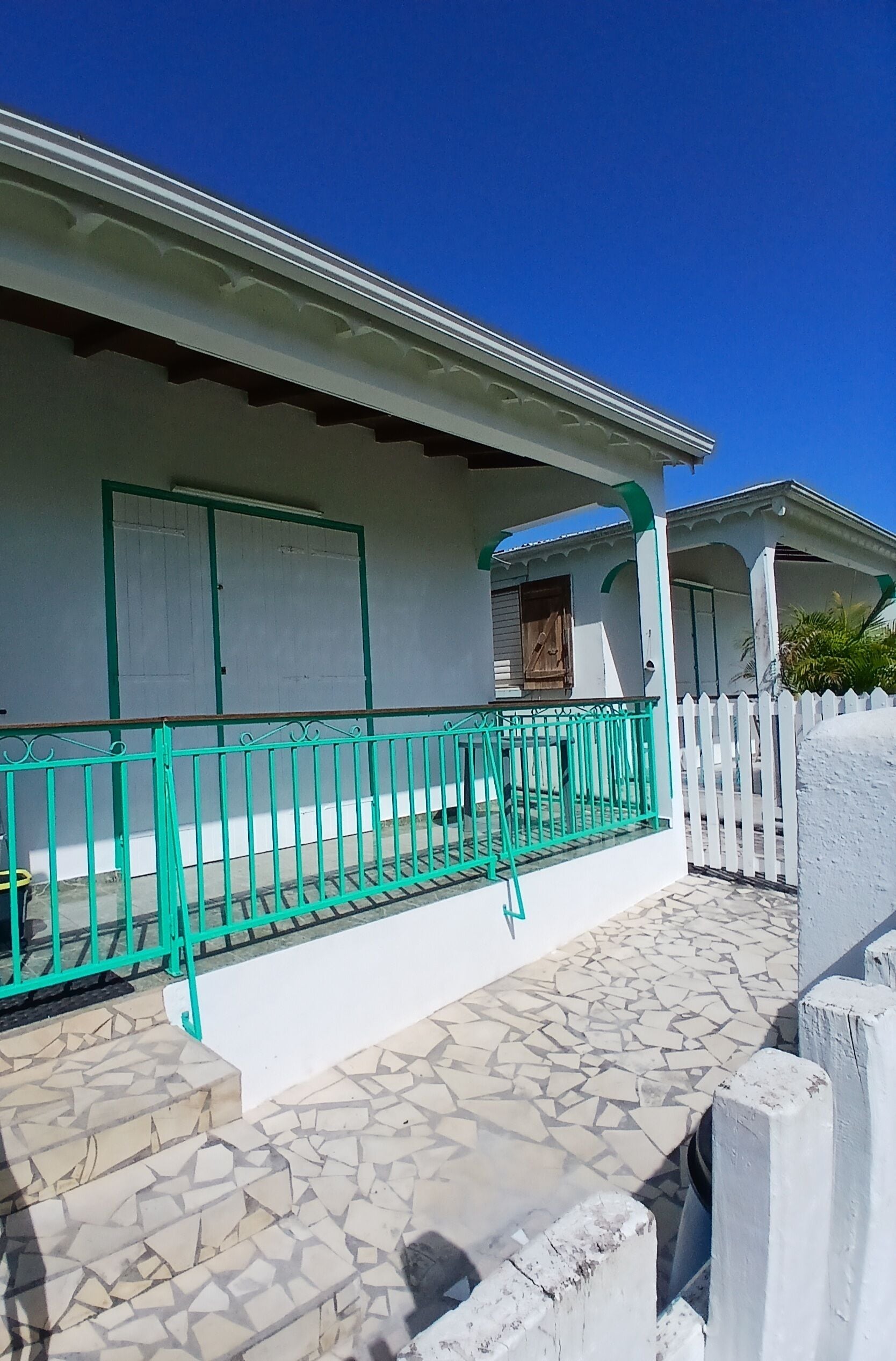 Une maison à l’extérieur blanc, avec une rambarde en métal vert et un porche carrelé en pierre.