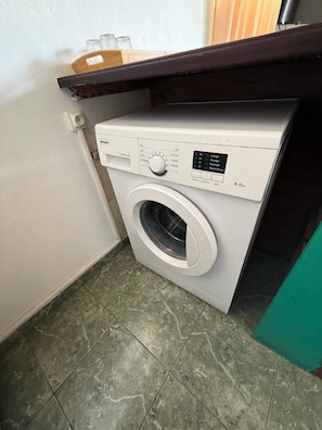 Una lavadora blanca de carga frontal y tamaño compacto, con panel de control, ubicada en un pequeño cuarto de lavado con piso de baldosas.
