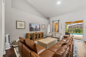 Ein geräumiges Wohnzimmer mit Ledersofas, einem Holzschrank, einem Flachbildfernseher und einem großen Fenster mit Blick auf einen Swimmingpool.