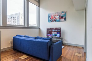 A room with a blue sofa, a flat-screen TV displaying a menu, and a window with a view of a building. - Downtown Loft Retreat - Nearby Parking - 2F (Louisville)