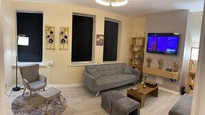 A modern living room with a grey sofa, a grey armchair, a small ottoman, a round rug, a flat-screen TV mounted on the wall, and a wooden shelf with decorative items. - Prestige 3 bedroom flat in Cardiff (Cardiff)