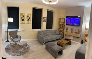 Ein modernes Wohnzimmer mit einem grauen Sofa, einem grauen Sessel, einem kleinen Hocker, einem runden Teppich, einem flachen Fernseher an der Wand und einem Holzregal mit Dekoration.