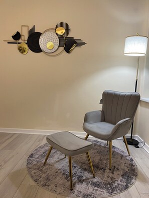 Ein Zimmer mit grauem Sessel und Hocker auf einem gemusterten Teppich, einer Stehlampe und einem Wandbild.