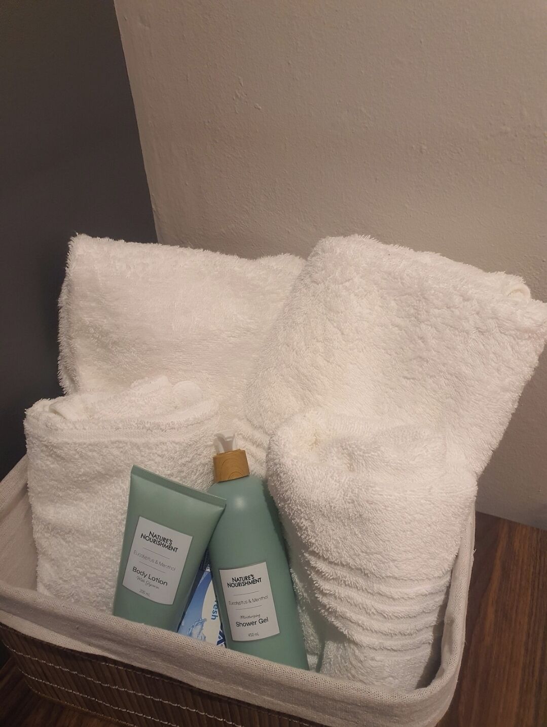 Une serviette blanche pliée, posée dans un panier, avec deux flacons de lotion corporelle et de gel douche de la marque Nature's Nourishment.