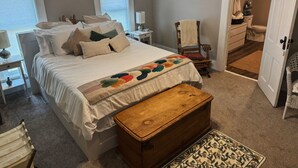 A bedroom with a bed, bedside tables, a wooden chest, a chair, and a bathroom visible through an open door.