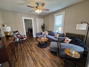A living room with a ceiling fan, a sofa set, a coffee table, a wooden floor, and a dining area in the background.