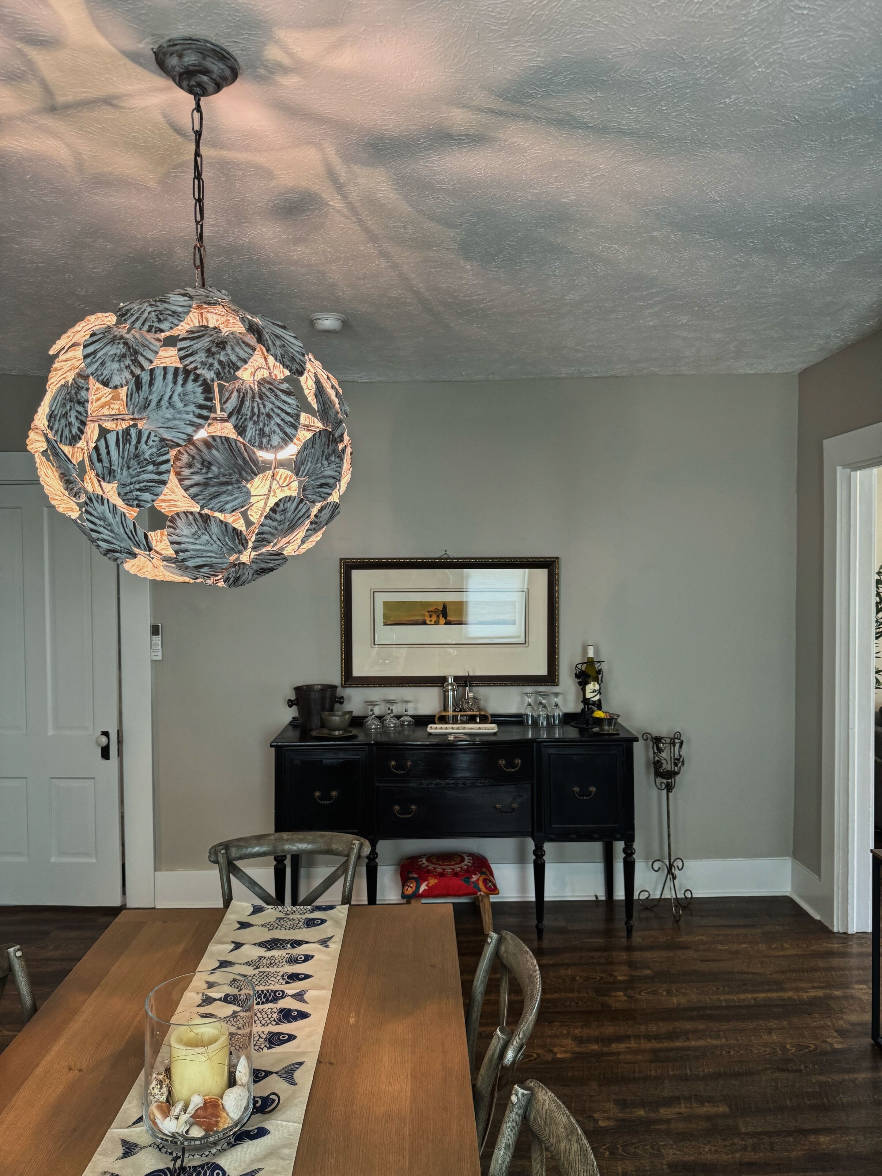 A dining area with a wooden table, chairs, a chandelier, a framed picture on the wall, and a dark wooden floor.