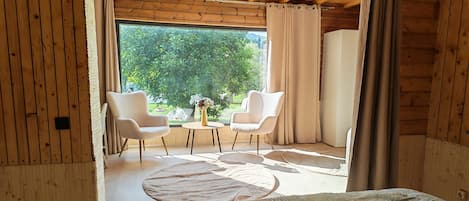 A room with a wooden ceiling, two chairs, a round table, a bed, and a view of greenery outside.