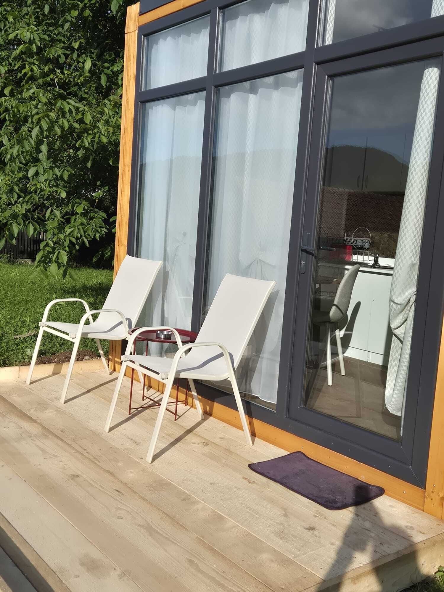 Una terraza con dos sillones blancos, piso de madera y puerta de vidrio que da a un espacio interior.