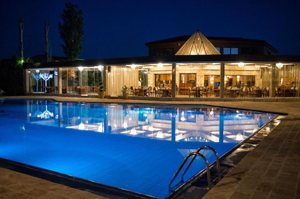A swimming pool with a glass enclosure, a dining area with tables and chairs, and a building with a triangular roof structure.