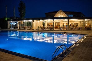 A swimming pool with a glass enclosure, a dining area with tables and chairs, and a building with a triangular roof structure. - CORALI RESORT (Agrinio)