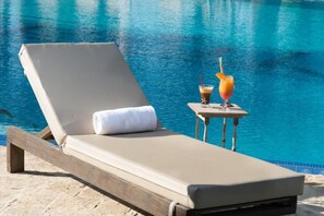 A sun lounger with a towel, a drink with a straw, and a table with a drink, all by a pool.
