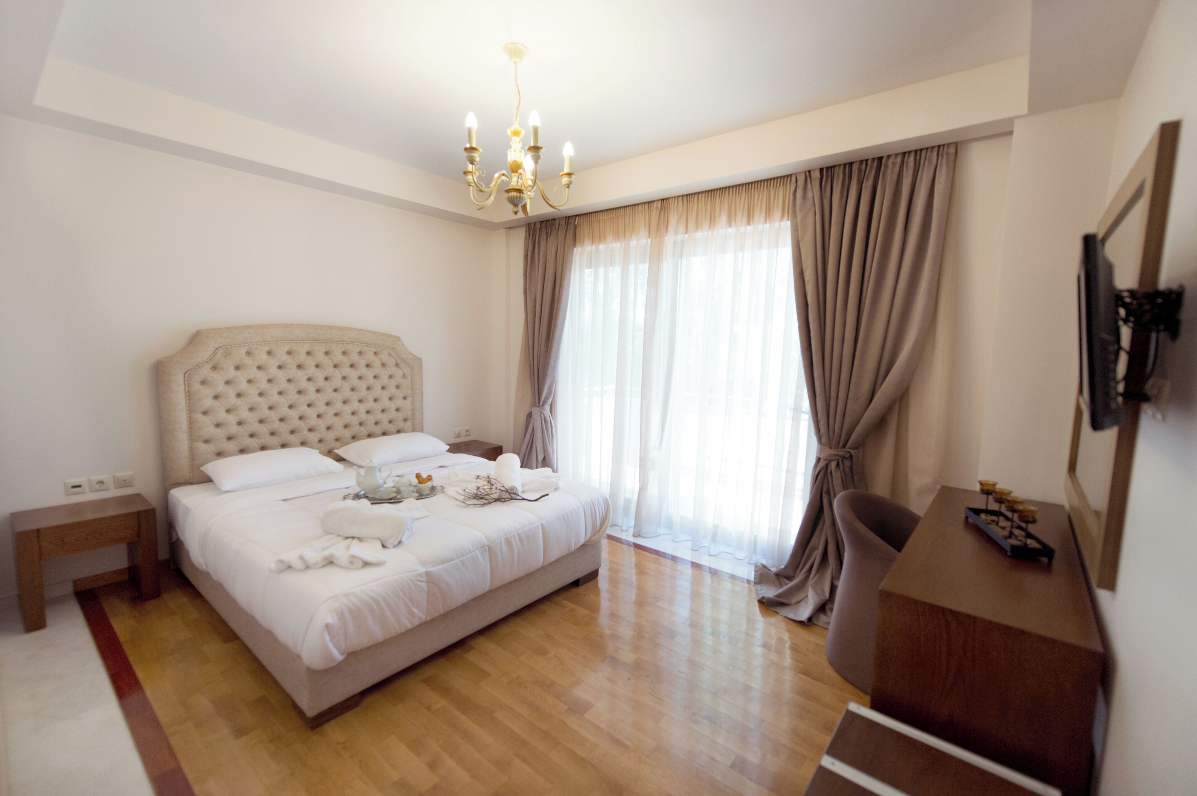 Habitación de hotel con una cama grande, piso de madera, candelabro, escritorio y televisor montado en la pared.