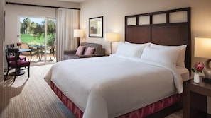 A hotel room with a large bed, a desk with a chair, a sofa, and a view of a golf course through a sliding door. - Coachella Rental for 2028 and 2029 (Palm Desert)
