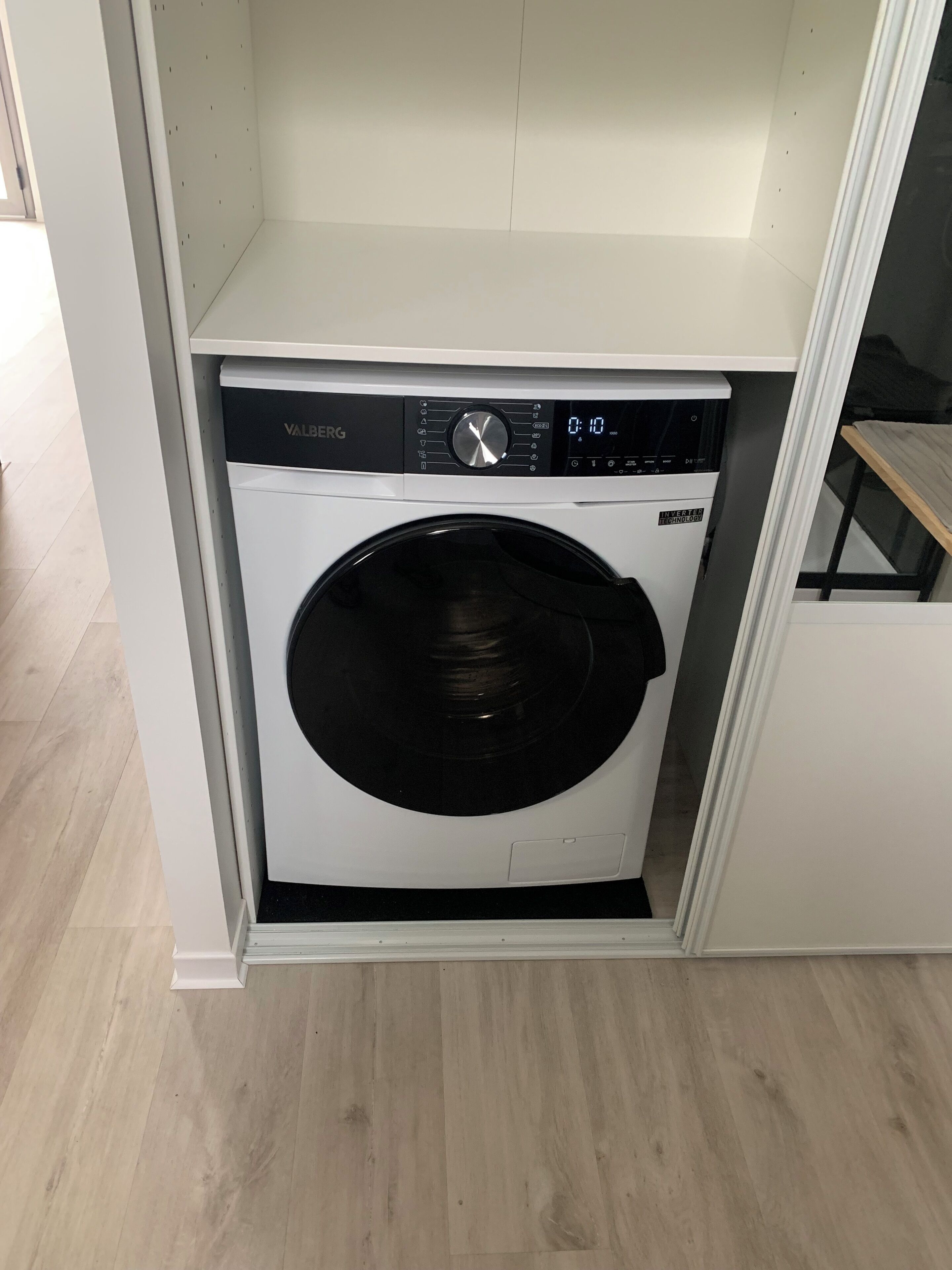 A compact, white front-loading washing machine in a small, enclosed laundry area with a wooden floor.
