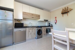 A modern kitchen with stainless steel appliances, wooden cabinets, a built-in washing machine, and a dining area with a white table and bench. - Casa Vista Mar - 2 Bed Apt With Sea Views & Communal Pool (Carvoeiro)