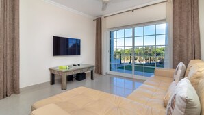 A living room with a sofa, a coffee table, a flat-screen TV, and a large window with a view of a pool and greenery. - Casa Vista Mar - 2 Bed Apt With Sea Views & Communal Pool (Carvoeiro)