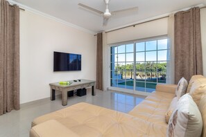 A living room with a sofa, a coffee table, a flat-screen TV, and a large window with a view of a pool and greenery. - Casa Vista Mar - 2 Bed Apt With Sea Views & Communal Pool (Carvoeiro)