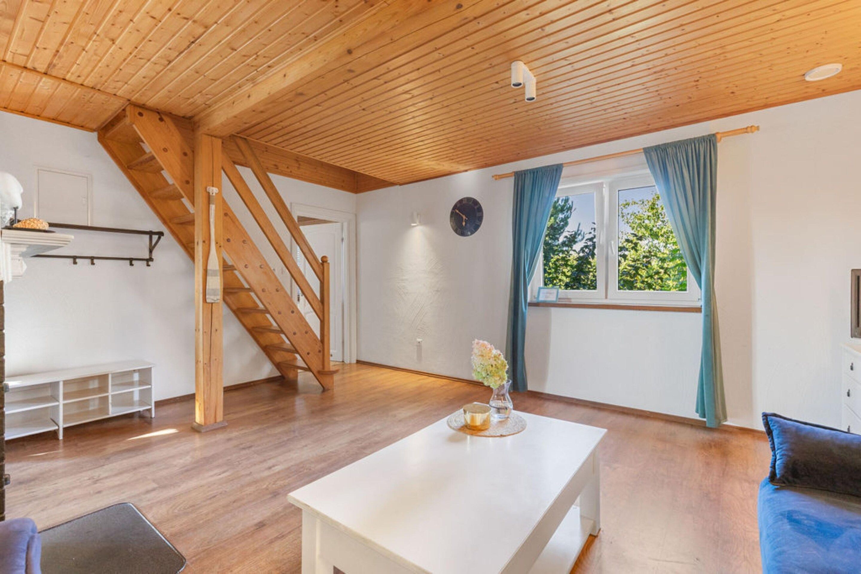 A room with a wooden staircase, a white table, a window with curtains, and a clock on the wall.