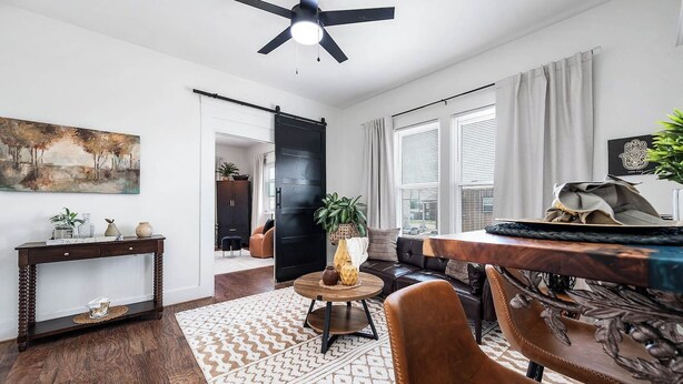 A modern living room with a dark wooden floor, a round wooden coffee table, a brown leather sofa, a dining table with a decorative centerpiece, and a ceiling fan. - Coastal Sophistication (Galveston)