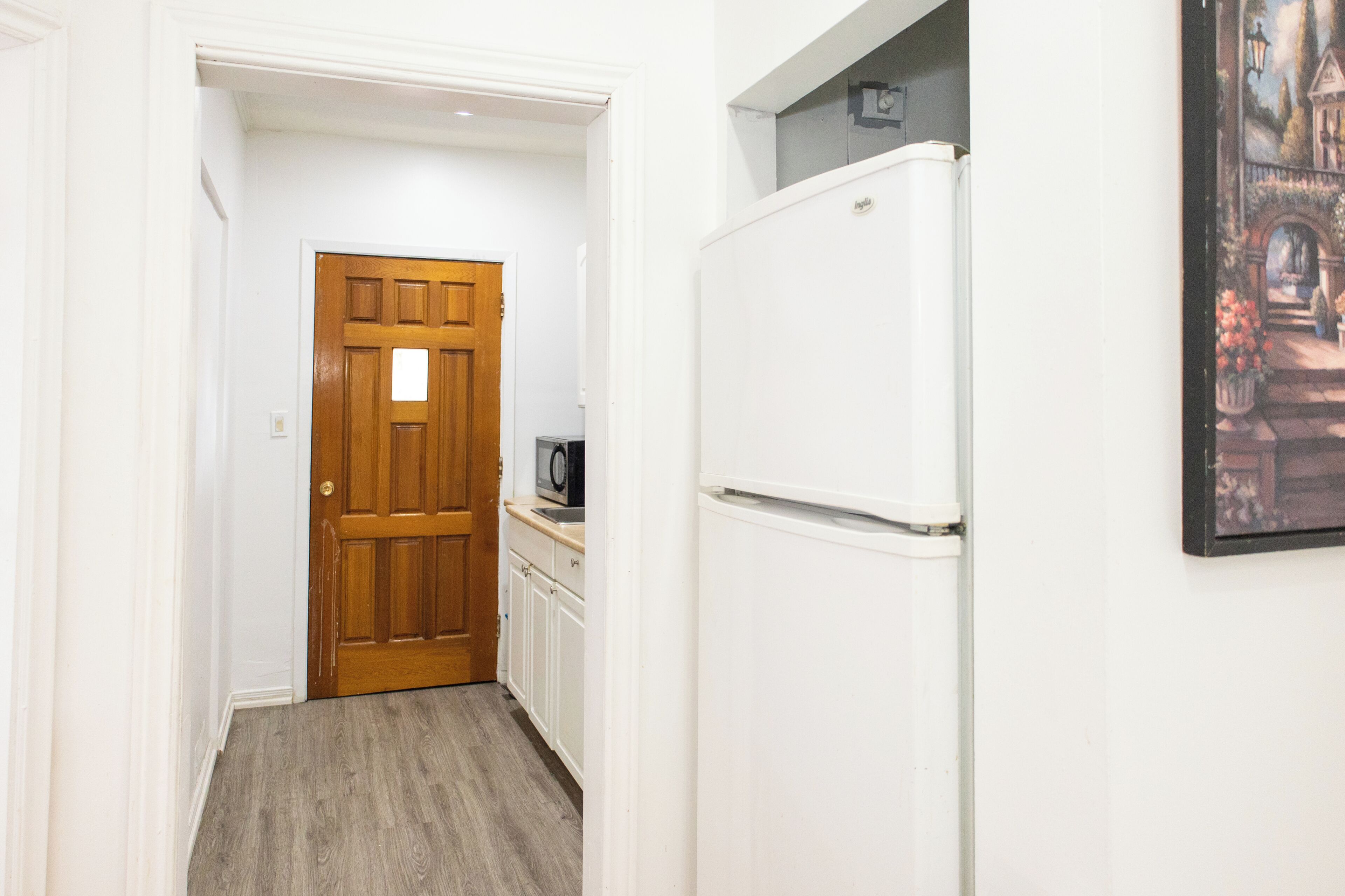 A narrow hallway with a wooden door, a white refrigerator, and a small kitchen area with a microwave.