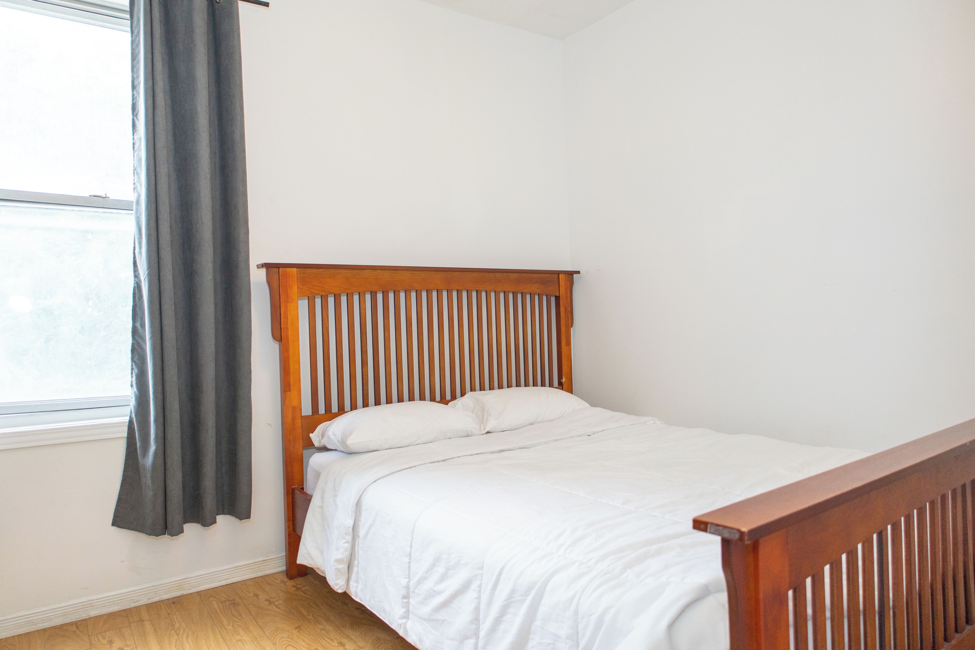 A single bed with a wooden headboard in a room with white walls and a window with a grey curtain.
