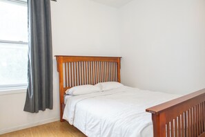 A single bed with a wooden headboard in a room with white walls and a window with a grey curtain. - Charming Stay in the heart of Chinatown. Explore and Have Fun (Ottawa)