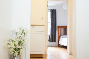 Un pasillo que conduce a un dormitorio con cama, una ventana con cortinas y un jarrón con flores.