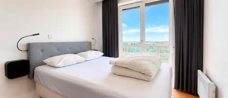A modern hotel room with a large bed, a bedside table, a window with curtains, and a view of the cityscape.